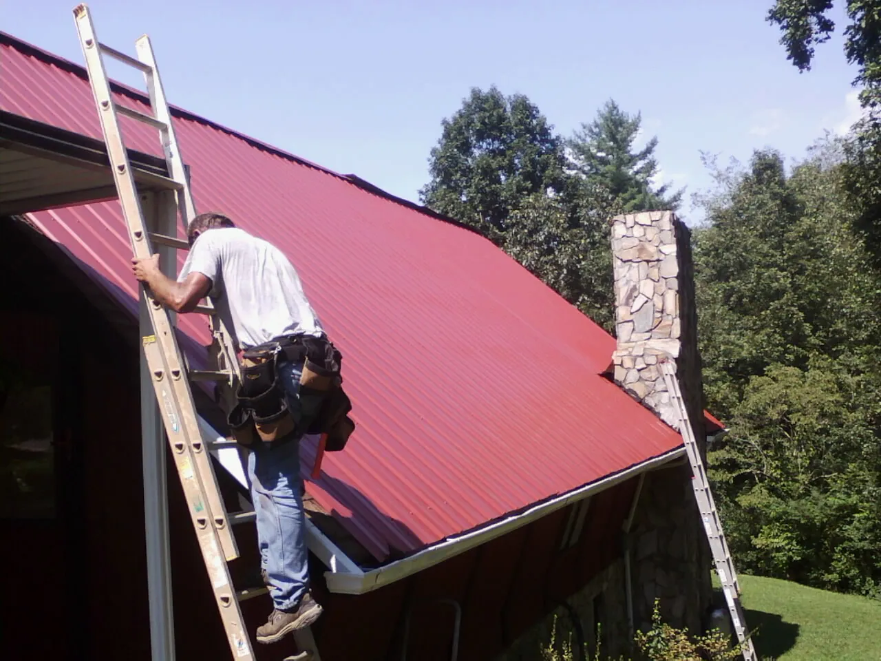 Roofer on a ladder beside a red metal roof and stone chimney — Soffit & Fascia Repair by Roofing Brothers in Wingate